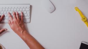 Hands typing on a keyboard with a watch and a cellphone to the side